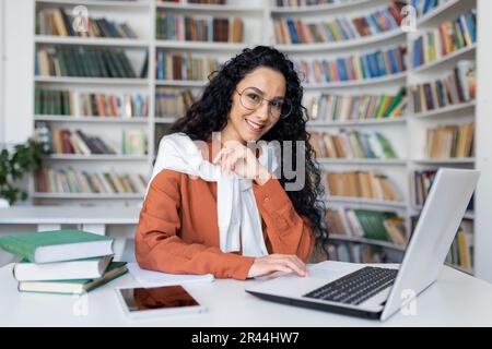 Jeune femme hispanique étudiant dans la bibliothèque universitaire, étudiante souriante et regardant l'appareil-photo tout en étant assise à un ordinateur portable, femme aux cheveux bouclés. Banque D'Images