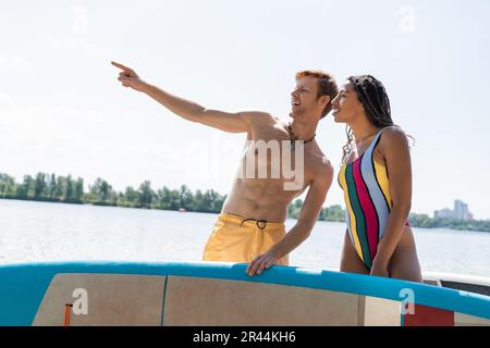 homme gai et rougehead regardant loin et pointant avec le doigt près de la femme afro-américaine dans un maillot de bain coloré tout en se tenant avec les planches sup près de la Banque D'Images