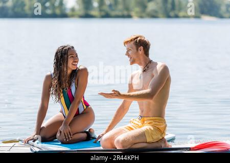 jeune homme à tête rouge pointant de la main et parlant à une femme afro-américaine gaie en maillot de bain coloré tout en étant assis sur des planches de sup sur l Banque D'Images