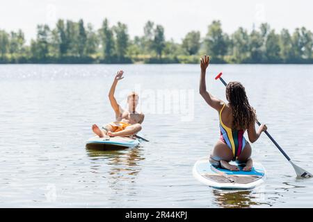 brunette afro-américaine femme en maillot de bain rayé et un homme à tête rouge surjoyeuse se faisant des mains en naviguant sur des planches de dessus sur le lac en été Banque D'Images