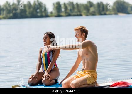un homme à tête rouge excité pointant du doigt près d'une femme afro-américaine stupéfaite en maillot de bain coloré tout en étant assis ensemble sur des planches de sup sur la rivière à summ Banque D'Images