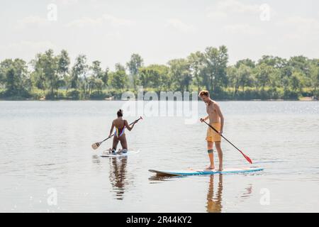 jeune et sportif redhead homme debout à bord de sup avec paddle près de la femme afro-américaine en maillot de bain coloré voile sur le lac pittoresque avec gre Banque D'Images