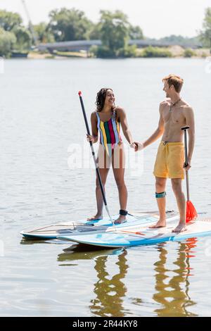 homme à tête rouge actif et femme afro-américaine joyeuse en maillot de bain rayé tenant les mains et souriant l'un à l'autre tout en se tenant debout sur des planches de sup sur lak Banque D'Images