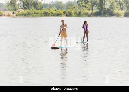 pleine longueur de jeune sportif homme et afro-américaine en maillot de bain rayé passer du temps sur le lac en naviguant sur des planches de sup avec des pagaies l'été Banque D'Images