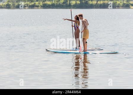 vue latérale d'une femme afro-américaine en maillot de bain coloré pointant avec la main près d'un jeune homme sportif en naviguant sur des planches de sup pendant le week-end sur le lac Banque D'Images