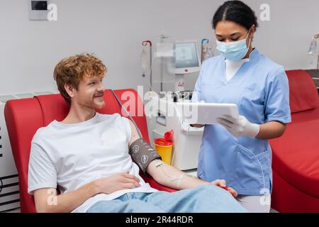 infirmier multiracial dans un masque médical et gants en latex utilisant un comprimé numérique près de la machine de transfusion et de la tête rouge donneur avec le brassard de pression artérielle sourire Banque D'Images