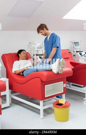 redhead médecin en uniforme bleu et gants en latex près des machines de transfusion automatisées et de la femme multiraciale sur une chaise médicale donnant du sang à l'hôpital, Banque D'Images