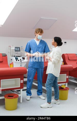 femme multiraciale parlant au médecin à tête rouge en uniforme bleu, masque médical et gants en latex près des chaises ergonomiques rouges et de la machine de transfusion automatisée Banque D'Images