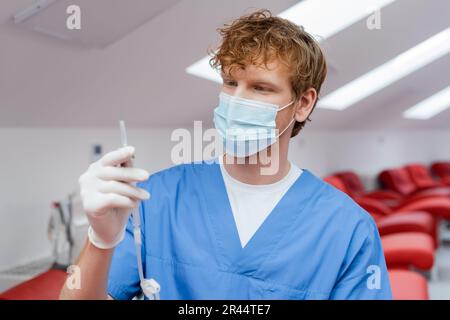 jeune et redhead médecin en uniforme bleu, masque médical et gant de latex tenant la transfusion sanguine ensemble près des chaises médicales ergonomiques sur le dos flou Banque D'Images