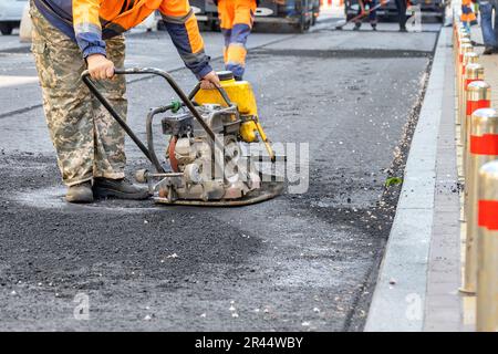 Un travailleur routier répare une section de route en compactant de l'asphalte frais avec une plaque vibrante autour d'un trou d'homme. Copier l'espace. Banque D'Images