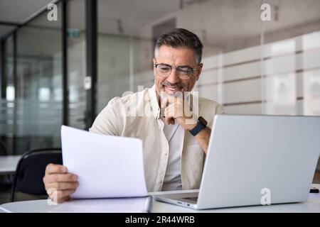 Homme d'affaires latin souriant analyste comptable tenant des documents, travailler à l'ordinateur portable Banque D'Images