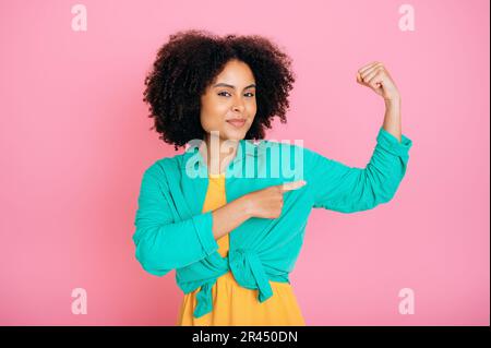 Positive belle femme brésilienne ou espagnole à la mode avec des cheveux bouclés, montrant des biceps sur son bras, montrant sa force, regardant l'appareil photo, souriant, se tenant sur un arrière-plan rose isolé Banque D'Images