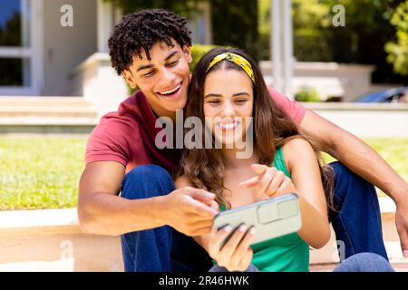 Un jeune couple biracial riant et prenant un selfie avec un téléphone portable tout en étant assis sur des marches dans la cour Banque D'Images