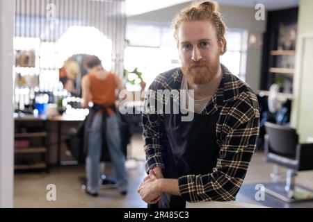 Portrait d'un coiffeur caucasien souriant barbu avec un petit pain en tablier noir au salon de coiffure Banque D'Images
