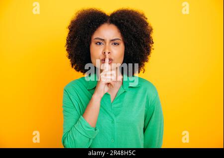 Secret, mystère. Photo d'une jolie femme brésilienne ou latino-américaine, vêtue d'une tenue décontractée, montrant le signe shh avec l'index touchant ses lèvres, arrête de parler, regarde l'appareil photo, se tient sur un fond jaune isolé Banque D'Images