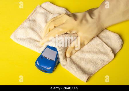 Une voiture à jouets bleue est nettoyée et désinfectée à la main recouverte d'un gant jaune et d'un chiffon en microfibre sur fond jaune Banque D'Images