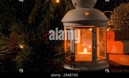 Lanterne blanche avec une bougie allumée. Livres, branche d'arbre de noël et lumières sur fond. Chute de neige. Image sombre de nuit. Humeur mystère de Noël Banque D'Images