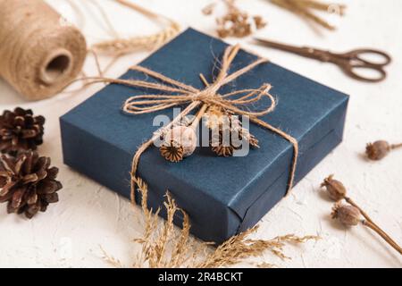 Boîte cadeau faite à la main dans un emballage bleu foncé, têtes de graines de pavot, fleurs séchées sur un bureau blanc. Cadeau pour homme. Fleuriste, herbier artisanal. Couleurs naturelles. Banque D'Images