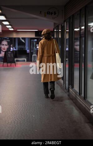 Une figure solitaire dans un manteau brun passe devant une fenêtre, avec un regard déterminé sur leur visage, dans un cadre urbain moderne Banque D'Images