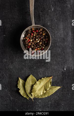 Grande vieille cuillère avec du poivre et des feuilles de Laurier séchées sur fond noir. Pose à plat. Vue de dessus. Concept alimentaire. Photographie de nourriture d'ambiance sombre. Banque D'Images