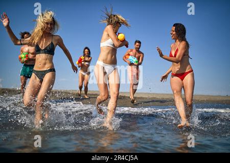 Jeunes hommes et femmes caucasiens jouant avec des pistolets à eau sur la rive. Groupe d'amis en vacances ensemble. Concept de convivialité, de plaisir, de style de vie. Banque D'Images