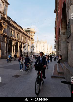 Homme portant un costume à vélo avec un panier à l'avant dans une rue de Milan avec le Duomo éclairé par la lumière du soir derrière, Lombardie, Italie Banque D'Images