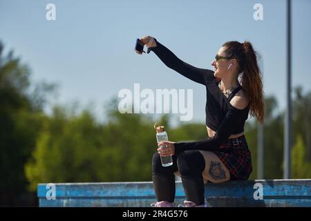 Joyeuse blaleur femelle prenant un selfie avec une bouteille d'eau en verre éco à la main. Portrait d'une personne patineuse assise sur une corniche dans un parc à roulettes et p Banque D'Images
