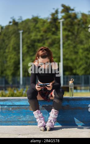 Patineuse en ligne assise sur une corniche dans un parc à roulettes et jouant à un jeu vidéo en ligne sur un smartphone avec une manette de jeu. Jeune rouleau froid blader personne g Banque D'Images