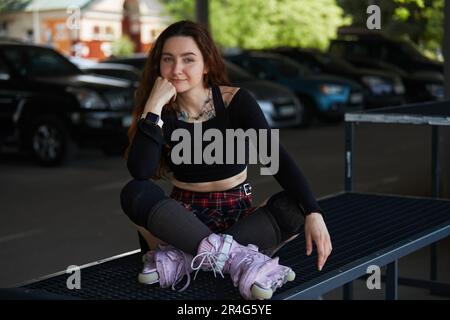 Patineuse en ligne assise dans un parc à roulettes avec des lames de rouleau agressives modernes sur ses pieds. Portrait d'une jolie jeune femme avec une ceinture thoracique tatouée Banque D'Images