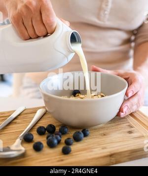 Une fille met du lait aux flocons d'avoine avec des myrtilles dans un bol pour un délicieux petit déjeuner de céréales dans la cuisine. Femme préparant du granola avec des baies et du latte pour un repas diététique du matin Banque D'Images