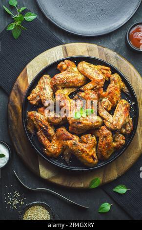 De délicieuses ailes de poulet à l'arrière-plan sont placées avec du sésame et de savoureuses sauces sur fond sombre. Vue de dessus avec gros plan. Banque D'Images