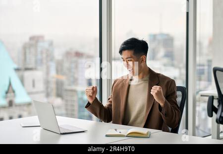Un homme d'affaires asiatique heureux qui célèbre son succès dans le travail au bureau en regardant un ordinateur portable. Banque D'Images