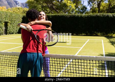 Jeune couple biracial avec raquettes de tennis embrassant tout en se tenant sur le court après le match Banque D'Images