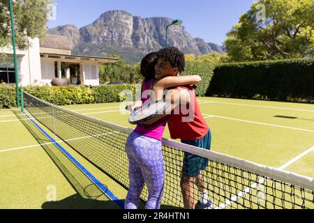 Jeune couple heureux biracial avec des raquettes embrassant tout en se tenant sur le court de tennis après le match Banque D'Images