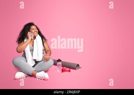 Femme noire surdimensionnée dans les vêtements de sport parlant sur le téléphone portable, assis avec des haltères et une bouteille d'eau, espace libre Banque D'Images