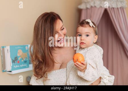 Jeune blonde, élégante mère tenant sa petite fille. Bébé tenant une orange Banque D'Images