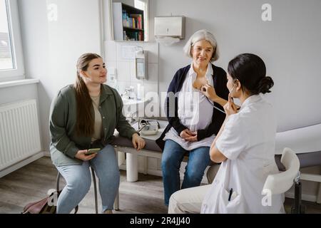 Un médecin écoute le rythme cardiaque d'une femme âgée assise avec sa fille à la clinique Banque D'Images