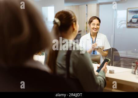 Une réceptionniste heureuse regarde la femme à travers une fenêtre de verre à l'hôpital Banque D'Images
