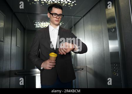 Jeune homme d'affaires avec une tasse de boisson chaude, heure d'enregistrement dans l'ascenseur Banque D'Images