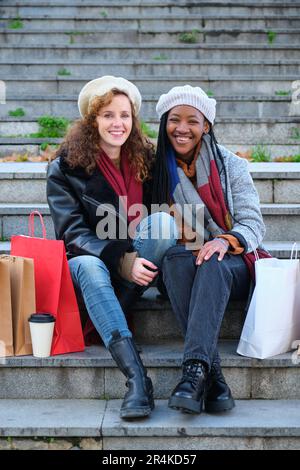 Deux amies multiraciales qui regardent l'appareil photo et sourient après avoir fait du shopping en hiver. Banque D'Images