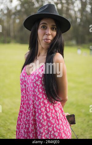 grimacing jeune femme avec de longs cheveux, porte chapeau et robe avec le modèle, en collant la langue pour le plaisir, le style de vie joyeux Banque D'Images