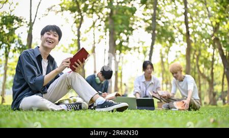 Un adolescent asiatique heureux et souriant étudiant dans des vêtements décontractés est assis sur l'herbe dans le parc du campus avec ses livres scolaires. Banque D'Images