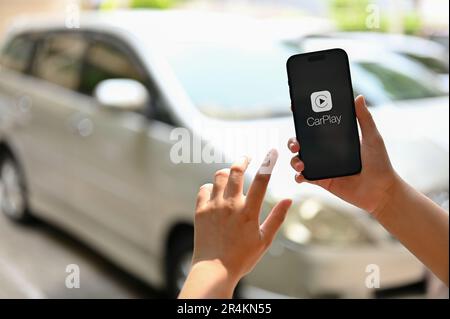 Chiang Mai, Thaïlande - 26 mai 2023: Gros plan d'une main femelle tenant un iPhone avec le logo CarPlay à l'écran sur un arrière-plan flou d'un c Banque D'Images