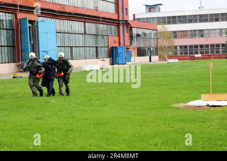 Trois pompiers secouristes en combinaisons de protection ignifuges et casques prennent la personne blessée dans un masque à gaz d'une zone dangereuse dans l'usine à nouveau Banque D'Images