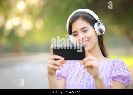 Une femme heureuse avec un casque regardant des vidéos au téléphone dans un parc Banque D'Images