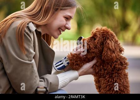 Une jeune fille blonde souriante dans un manteau de pluie beige élégant et un Jean joue avec son animal de compagnie jouet coodle tout en marchant dans un beau parc. Gros plan. Banque D'Images