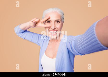 Une dame âgée fait un selfie sur l'appareil photo d'un smartphone faisant un v-sign près des yeux isolés sur fond violet clair avec un espace de copie pour le texte Banque D'Images