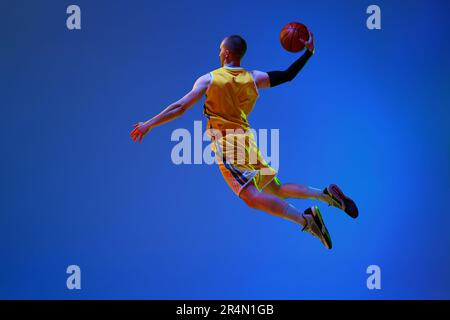 Image de la vue arrière d'un jeune joueur de basket-ball masculin en uniforme jaune, sautant avec le ballon sur fond bleu de studio à la lumière du néon. Banque D'Images