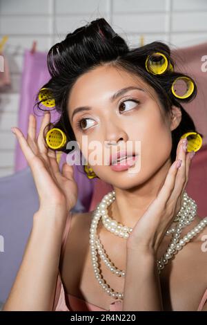 jeune femme asiatique pensive touchant les cheveux avec les cheveux curlers et posant dans collier de perles près de linge propre et humide accroché sur un arrière-plan flou, maison Banque D'Images