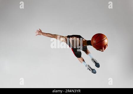 Sportif motivé, joueur de basket-ball masculin en mouvement, sautant avec le ballon sur fond gris de studio. Vue de dessus Banque D'Images